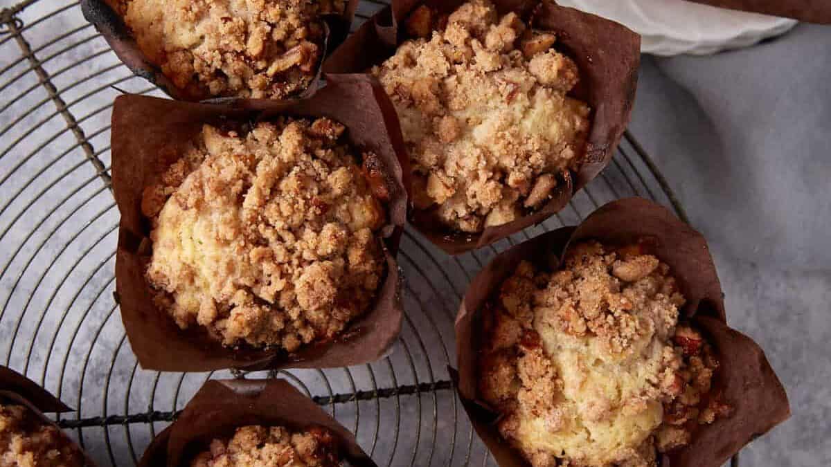 Top view of eight freshly baked muffins with crumbly tops in brown paper liners, arranged on a cooling rack. One muffin is set apart on a white plate, its liner partially unwrapped. A light gray napkin is casually draped in the background.