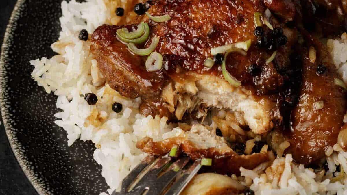 A close-up of a plate of white rice topped with glazed chicken, garnished with sliced green onions and whole black peppercorns—an easy option for affordable dinners. A fork is pulling apart a piece of the chicken.