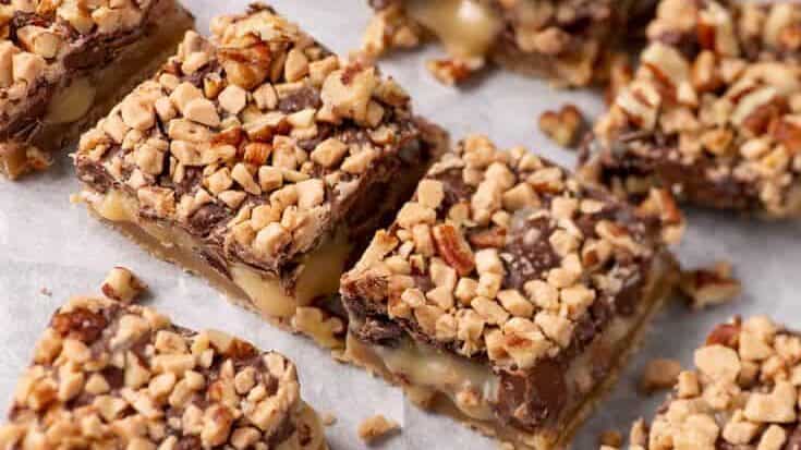 A batch of caramel and chocolate dessert bars made with sweetened condensed milk is arranged on a piece of parchment paper. The bars are cut into square pieces, showcasing layers of caramel and chocolate beneath the chopped nut topping.
