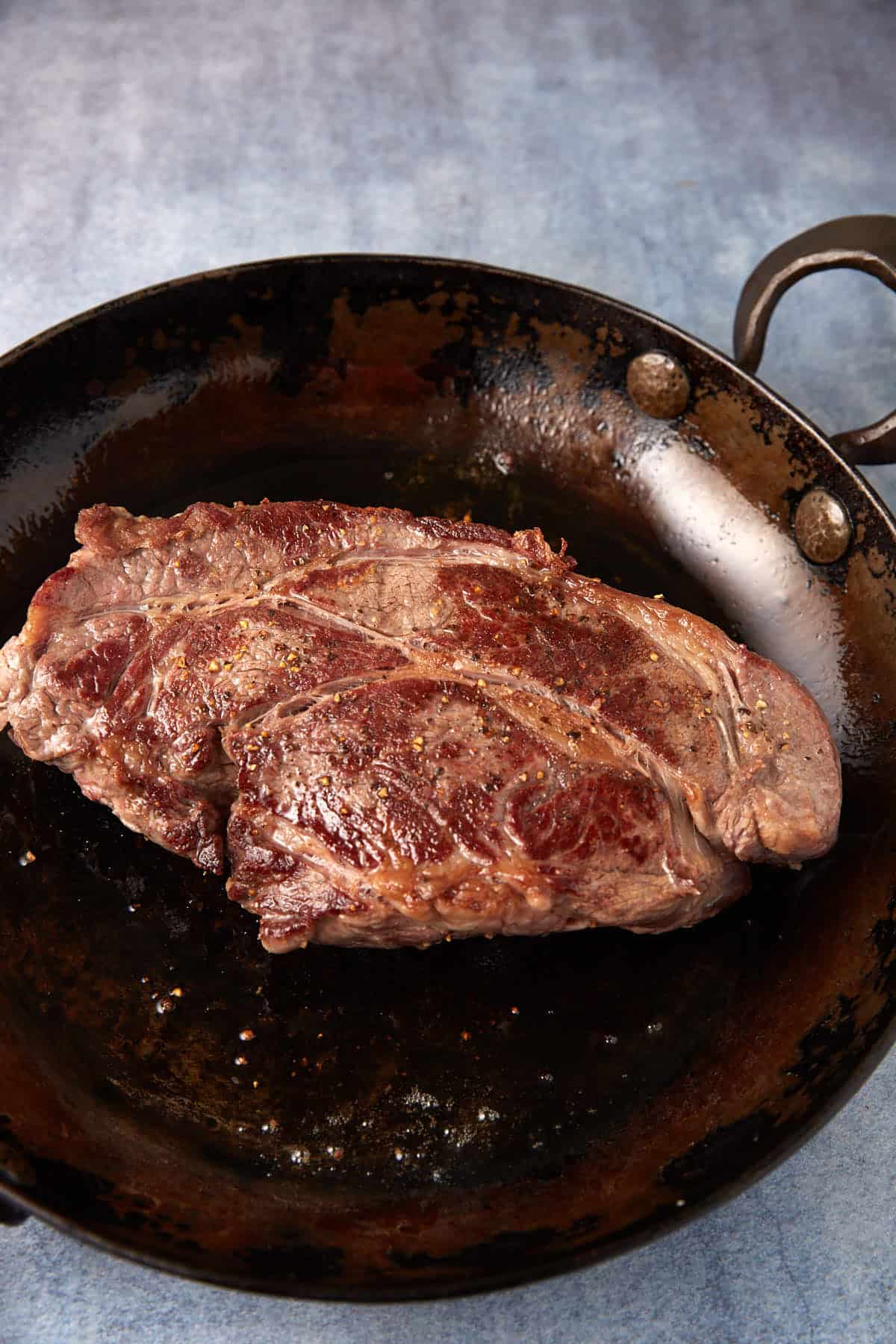 A seared, medium-rare steak sits in an old, well-seasoned cast-iron skillet. The steak is browned with visible grill marks and seasoned with salt and pepper. The background evokes a feeling similar to that of a slow cooker pot roast with gravy on a cold day against a blue slate surface.