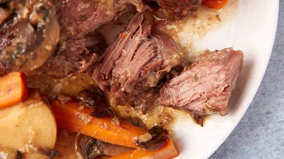 A close-up of a plate of slow cooker pot roast with gravy, featuring tender, shredded beef, carrots, mushrooms, and potatoes in a savory sauce. The dish is placed on a white plate against a light blue tablecloth background.