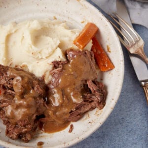 A plate of food featuring mashed potatoes, slow cooker pot roast with gravy, and cooked carrots. The meal is served on a white ceramic dish and placed on a blue table surface. A fork and knife rest beside the plate.