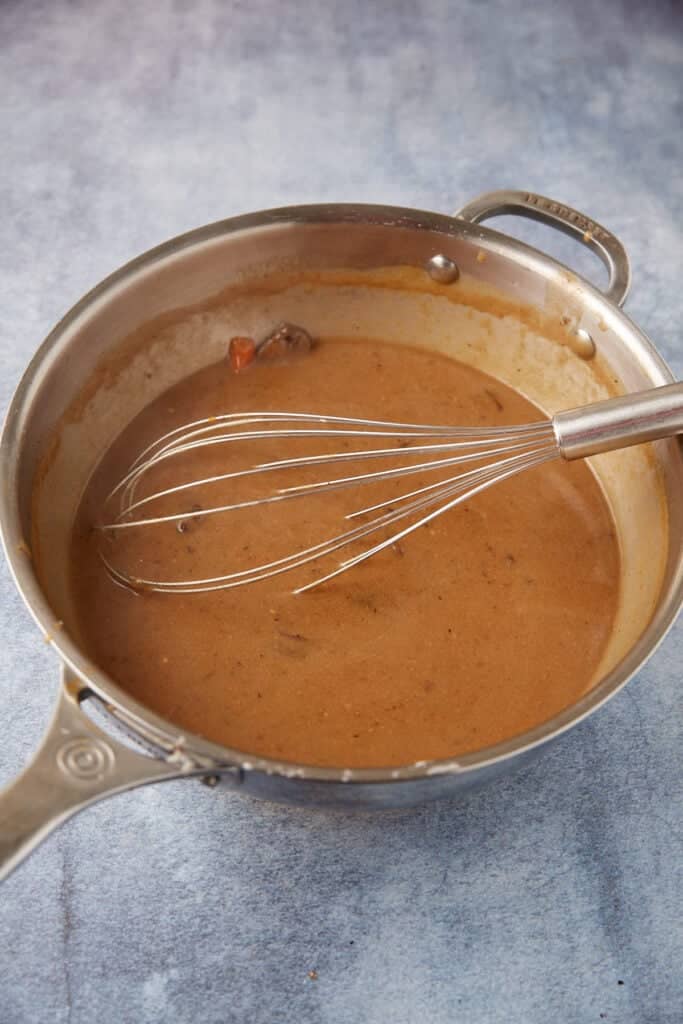 A metal whisk rests in a stainless steel saucepan filled with rich, brown gravy. The saucepan is placed on a blue-gray textured surface, reminiscent of preparing slow cooker pot roast with gravy.