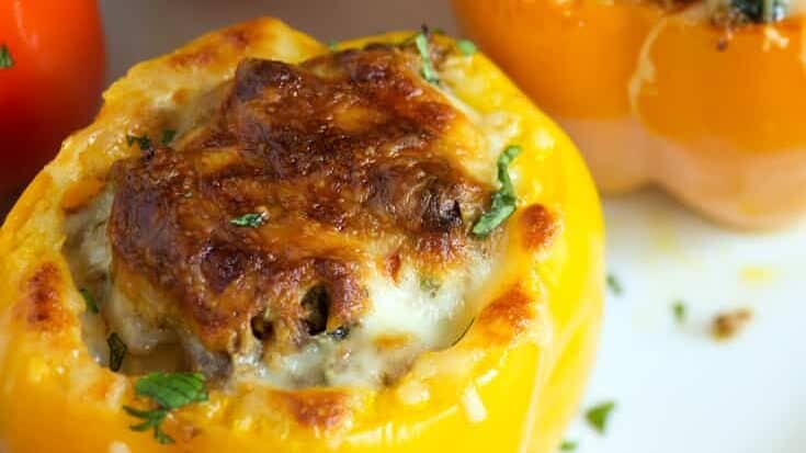 Four stuffed bell peppers, two yellow and two red, are filled with a savory mixture and baked with a golden brown, melted cheese topping. They are garnished with chopped herbs. The background features some green leafy vegetables. The dish is presented on a white plate.