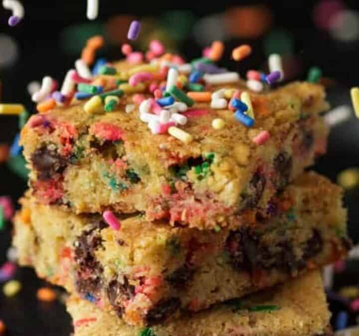 Three colorful funfetti blondies with chocolate chips, made in an 8x8 pan, are stacked on top of each other and surrounded by rainbow sprinkles against a dark background.
