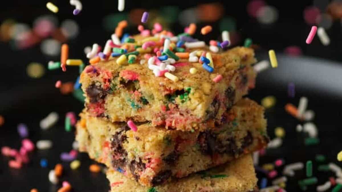 Three colorful funfetti blondies with chocolate chips, made in an 8x8 pan, are stacked on top of each other and surrounded by rainbow sprinkles against a dark background.