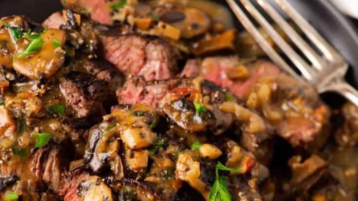 A plate of Steak Marsala topped with a rich mushroom sauce, garnished with chopped parsley. A fork rests on the side of the dish. The steak is sliced, revealing a medium-rare interior. The overall presentation is appetizing and savory.