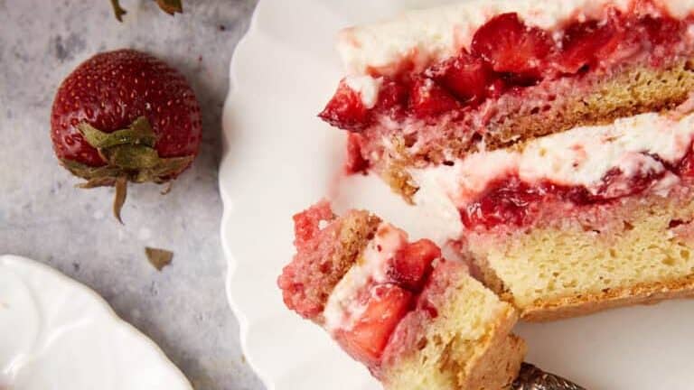 A slice of strawberry shortcake on a white scalloped plate, with a fork holding a piece of the cake. Two fresh strawberries are placed beside the plate. Another smaller plate with a piece of the shortcake is partially visible in the lower left corner.