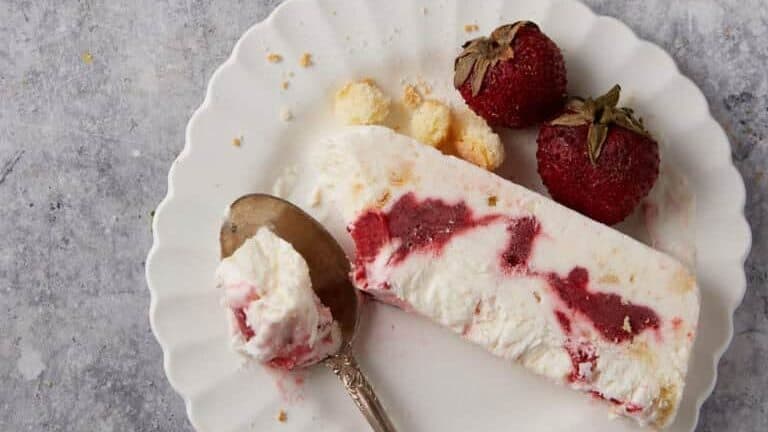 A dessert plate featuring a slice of creamy strawberry swirl dessert is accompanied by two fresh strawberries and a spoon. Another plate and tray with additional slices and spoons are in the background on a gray stone surface.