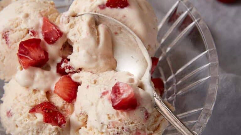 A glass bowl filled with three scoops of strawberry ice cream, garnished with fresh strawberry chunks. A spoon rests in the bowl, positioned as if about to scoop some ice cream. A few whole strawberries are scattered in the background on a gray surface.