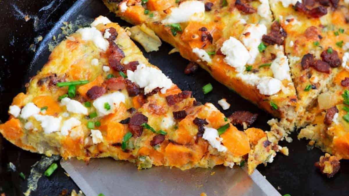 A delicious slice of Sweet Potato Frittata featuring chunks of sweet potatoes, crumbles of goat cheese, bits of crispy bacon, and a sprinkle of fresh green chives, all presented in a black skillet with a spatula lifting one slice.