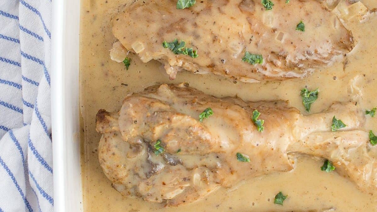 A close-up of a white baking dish containing several cooked chicken drumsticks covered in a creamy, brown sauce. The dish is garnished with small green herbs. A blue and white striped cloth is partially visible beside the dish.
