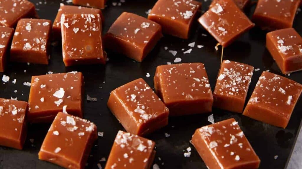 Close-up of several pieces of homemade salted caramel fudge, a perfect treat for caramel desserts lovers, topped with flaky sea salt and arranged on a dark surface.