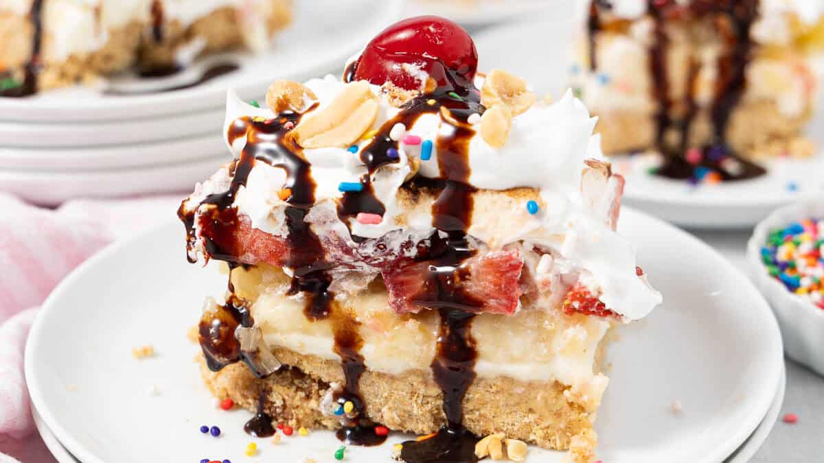 A slice of banana split dessert, one of the best easy desserts, is topped with whipped cream, chocolate syrup, chopped nuts, colorful sprinkles, and a cherry on a white plate. Other dessert slices and sprinkles are visible in the background.