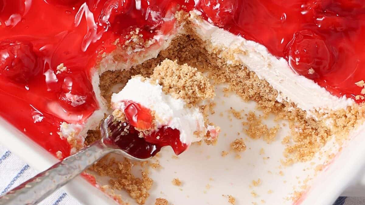 A close-up of a cream cheese dessert with a graham cracker crust, creamy white filling, and cherry topping. A spoon has scooped out a portion, revealing the luscious layers beneath.