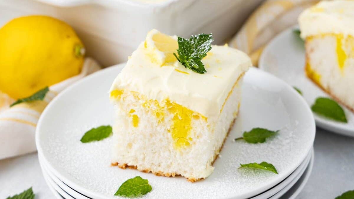 A square piece of lemon poke cake with frosting and mint leaves on top sits on a stack of white plates. The background shows a baking dish with the remainder of the cake, additional mint leaves, and a whole lemon.