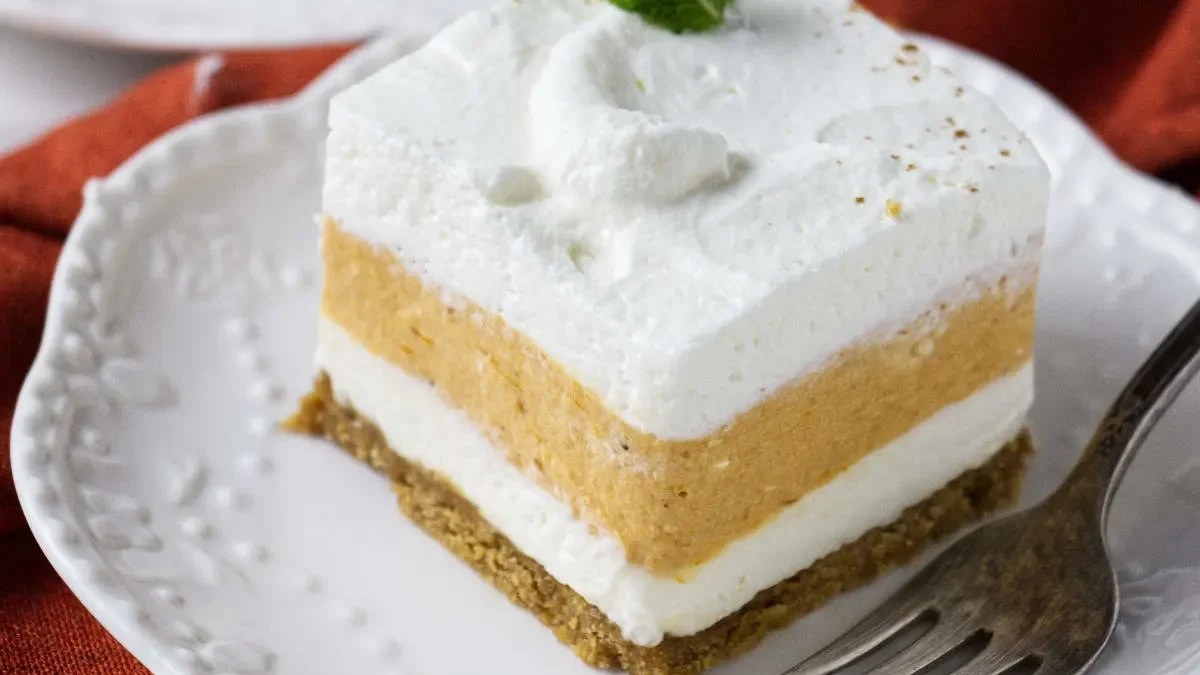 A delectable layered pumpkin dessert on a white plate with a fork. The treat features a creamy white base, an orange middle layer, and a whipped cream topping, garnished with a small mint leaf. In the background, more servings of the pumpkin desserts are on plates over a red cloth.