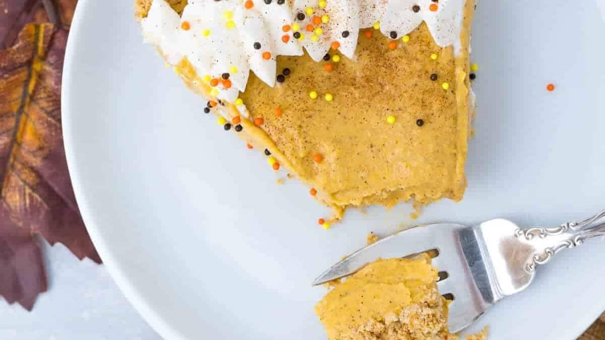 A slice of pumpkin pie topped with whipped cream and festive sprinkles sits on a white plate. A fork with a bite of pie rests on the plate. Autumnal leaves and a whole pumpkin pie in a tin are visible in the background.