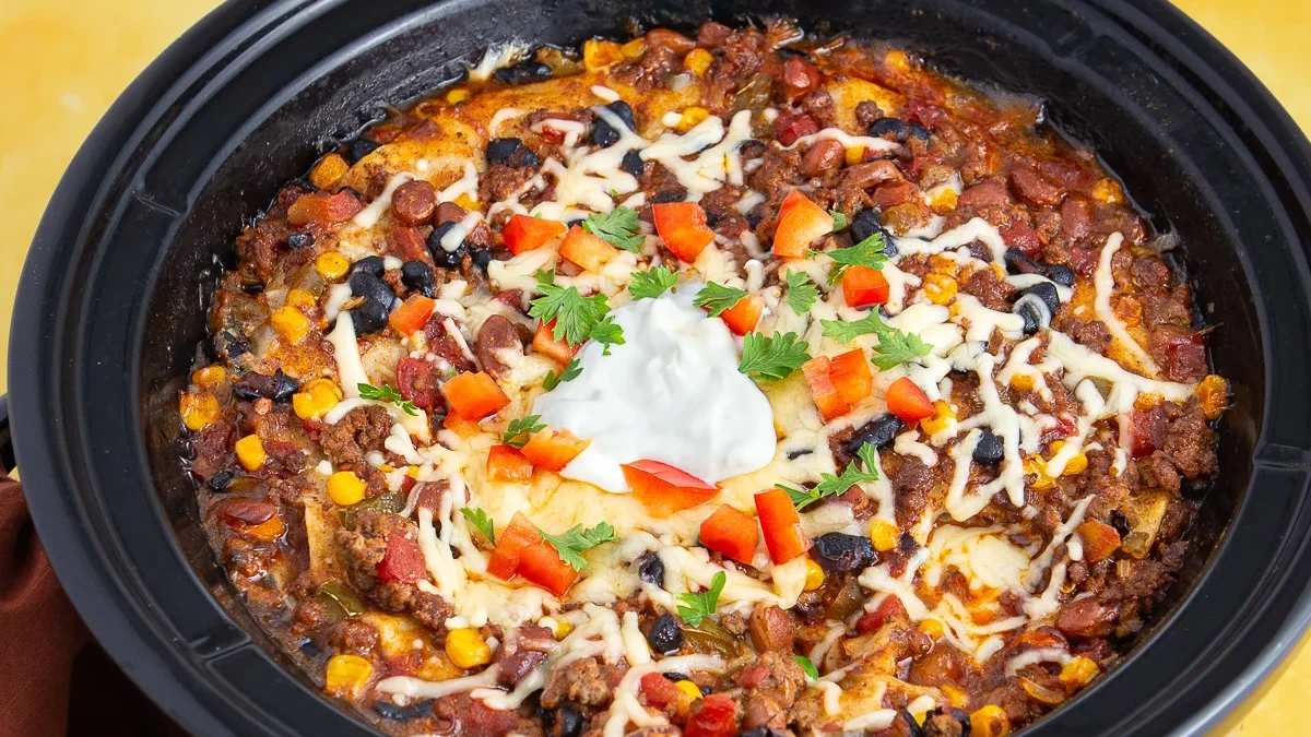 A slow cooker filled with chili made from one pound ground beef, topped with melted cheese, sour cream, chopped red bell peppers, and fresh parsley. Visible ingredients include beans, corn, and tomatoes.