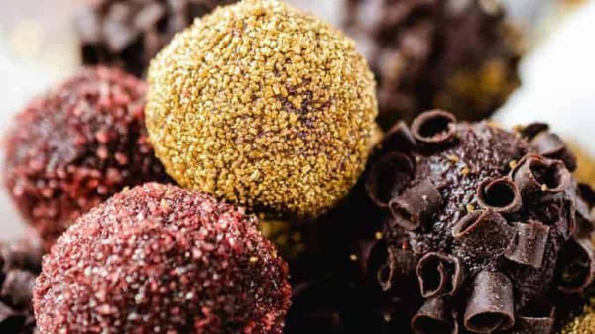 A close-up shot shows an assortment of beautifully decorated truffles. Some are coated in gold and red sparkly sugar, while others are covered in dark chocolate curls. The background features festive, curly ribbon decorations.