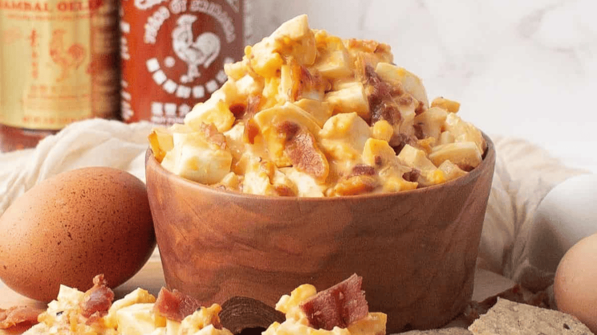 A wooden bowl filled with a chunky egg salad mixed with bacon pieces sits on a light surface, surrounded by whole eggs, a sauce bottle, and bread slices in the background—perfect for your Thanksgiving salad recipes collection.