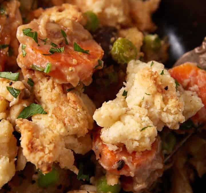A close-up of a black bowl filled with seasoned chicken and vegetable casserole. The dish includes chunks of bread or biscuit-like pieces, carrots, green peas, and chicken, all garnished with fresh herbs. A fork is partially visible, resting on the rim of the bowl.