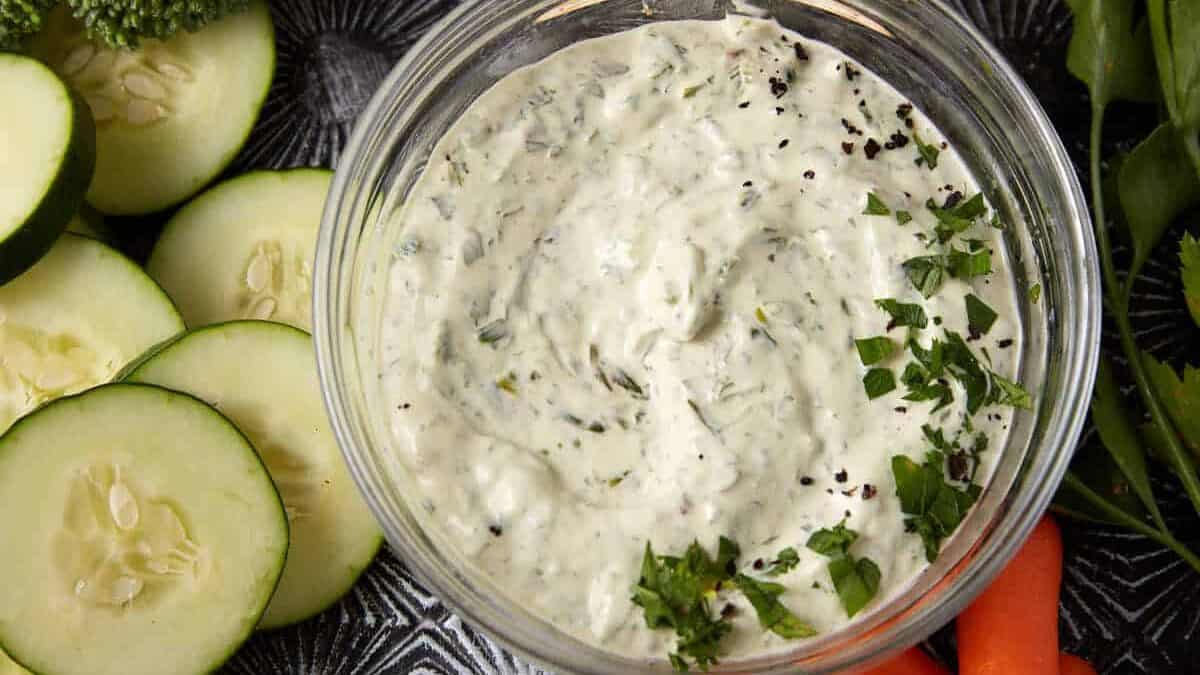 A bowl of creamy herb dip is surrounded by fresh vegetables, including broccoli, cucumber slices, baby carrots, celery sticks, and round crackers. The dip is garnished with chopped herbs and black pepper. The background features a textured, dark surface.