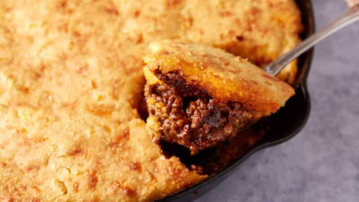 A close-up of a skillet filled with tamale pie, a delicious take on beef casseroles. A serving spoon lifts out a portion, revealing seasoned ground beef filling beneath a golden, baked cornbread crust.