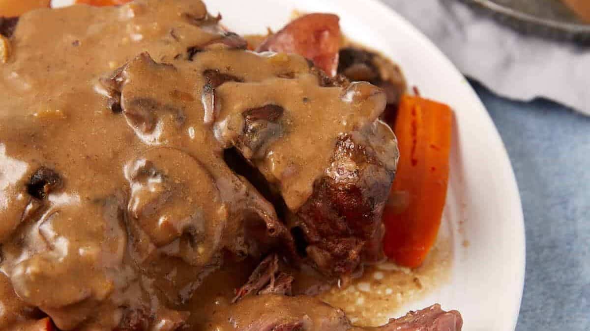 A plate of slow cooker pot roast with gravy, garnished with carrots, mushrooms, and potato chunks. A small dish of extra gravy lies in the background on a gray napkin. The meal is served on a white plate against a light blue surface.