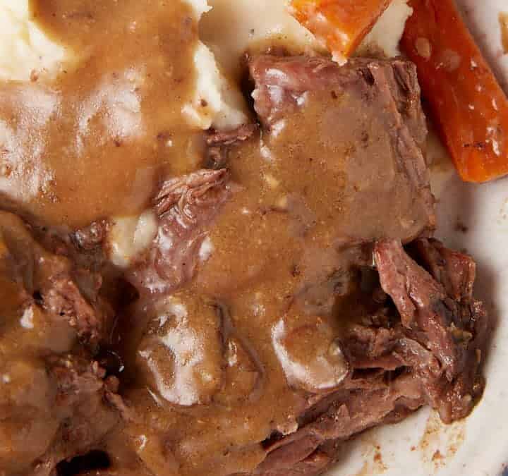 A plate of mashed potatoes topped with brown gravy, accompanied by slow-cooked beef and roasted carrot sticks. There is a fork and knife on the right side of the plate, and a metal jug of gravy in the background on a cloth napkin. This hearty meal features savory slow cooker pot roast with gravy.