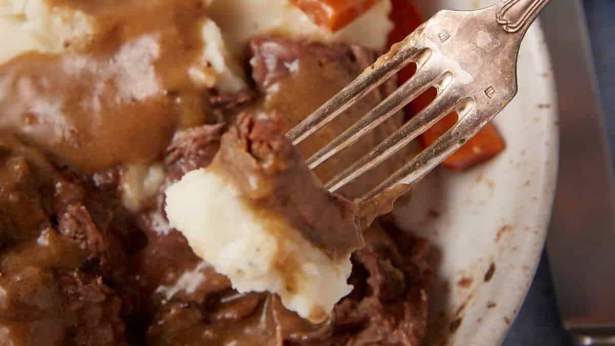 A close-up of a fork scooping mashed potatoes and gravy from a white ceramic bowl, filled with tender chunks of slow cooker pot roast with gravy and pieces of carrots. A knife rests beside the bowl, while a brown block of cheese appears in the background.