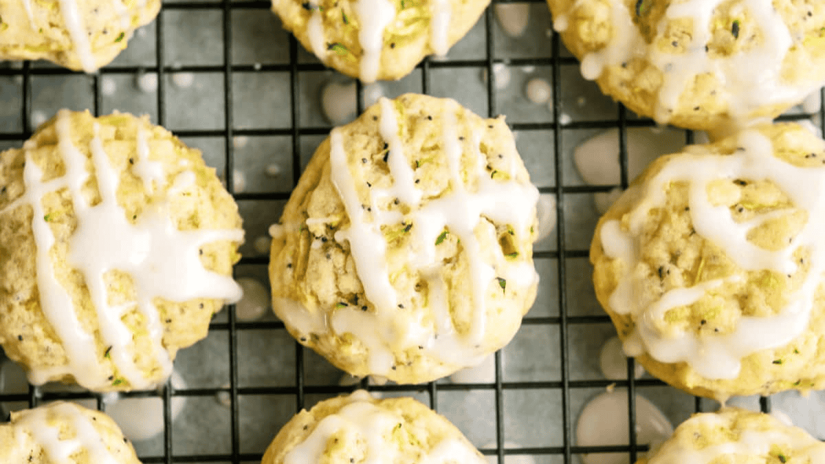 Round cookies with a light drizzle of white icing are cooling on a black wire rack. The cookies appear soft and have small green specks, making them a refreshing choice for fans of lemon desserts.