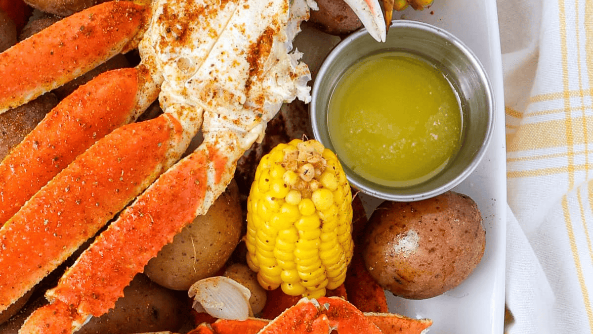 A plate featuring crab legs, corn on the cob, red potatoes, garlic cloves, and a metal cup of melted butter makes this a standout among one pot dinner recipes, with a yellow and white striped cloth nearby.