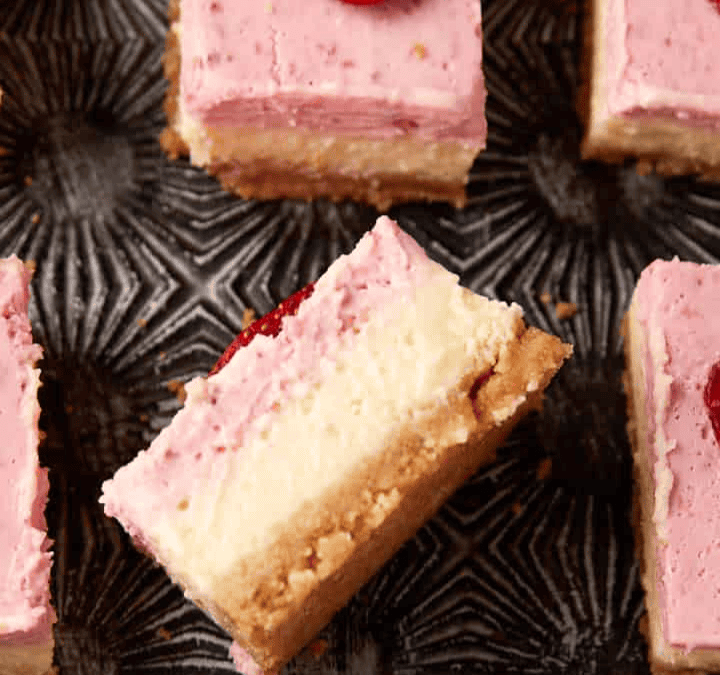 Several square strawberry cheesecake dessert bars with pink topping, crumbly crust, and strawberry slices on top are arranged on a dark patterned surface. One bar is turned to show its creamy layers.