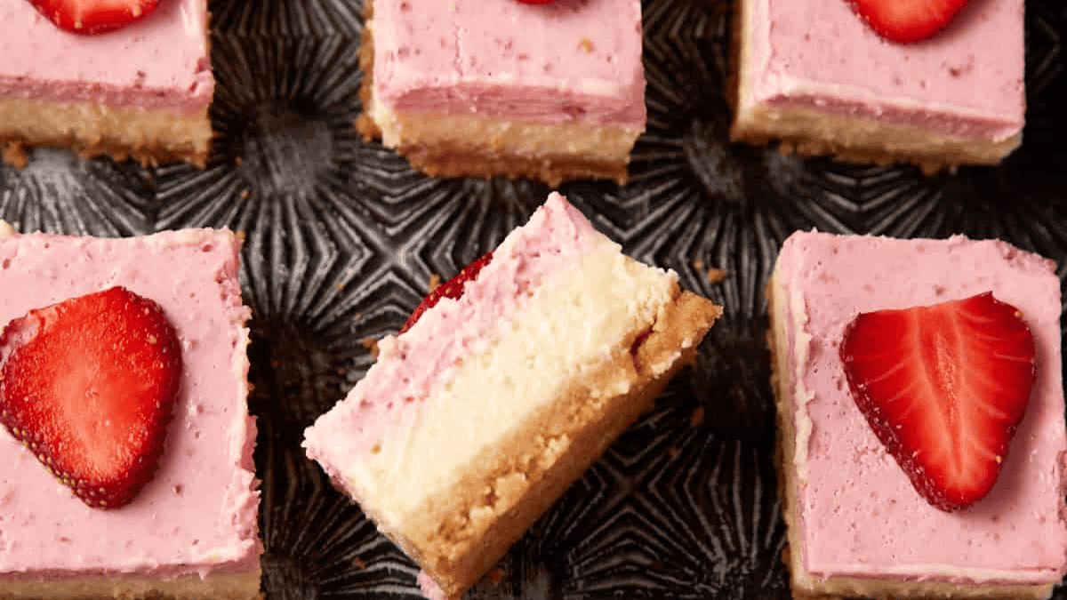Several square strawberry cheesecake dessert bars with pink topping, crumbly crust, and strawberry slices on top are arranged on a dark patterned surface. One bar is turned to show its creamy layers.