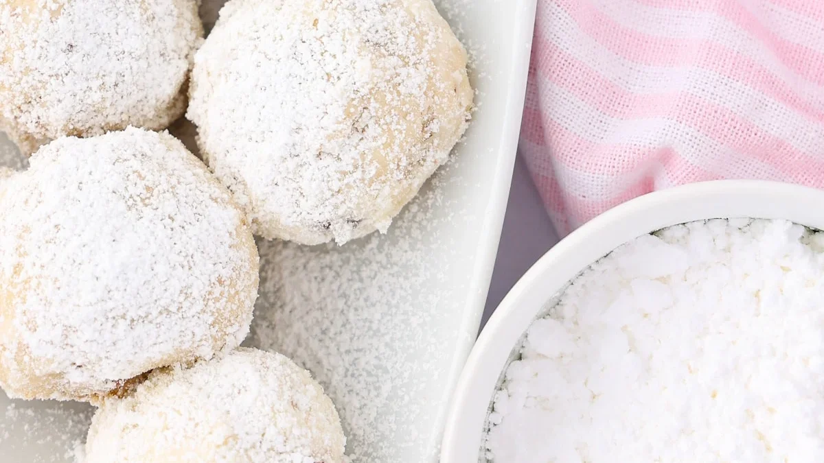 Powdered sugar cookies arranged on a white plate next to a bowl of powdered sugar and a pink-and-white striped cloth—classic treats inspired by retro dessert recipes.