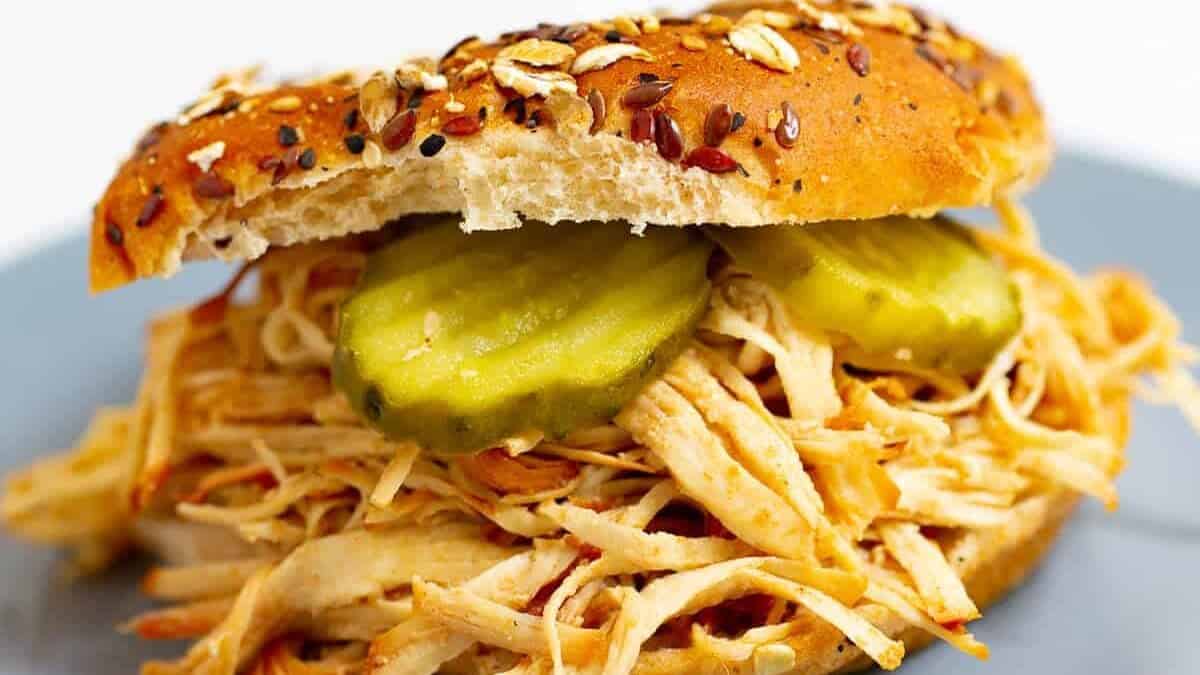A close-up of a shredded chicken sandwich with pickles on a multigrain bun, served on a gray plate—perfect for game day potluck recipes.