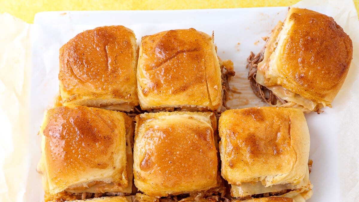 Six baked slider sandwiches with golden-brown tops are arranged closely on parchment paper. Perfect for game day potluck recipes, these sliders are stuffed with shredded meat and cheese, and the shiny tops look brushed with butter.