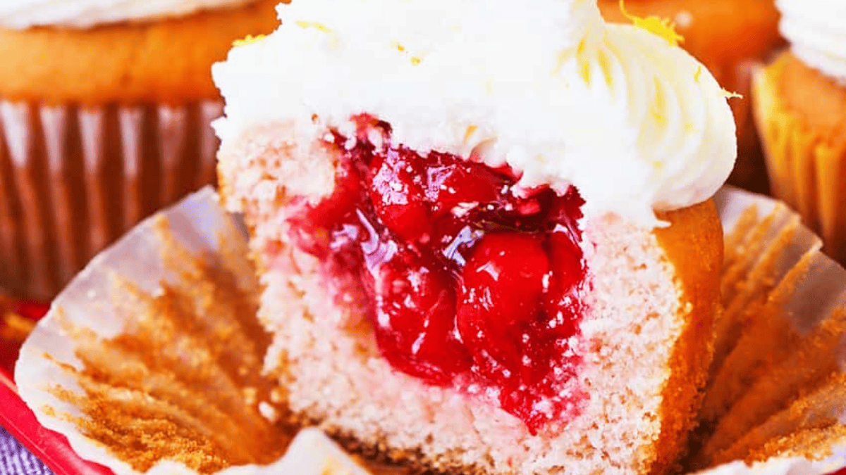 A close-up of a cupcake with a bite taken out, revealing a cherry filling inside. Perfect for holiday cherry desserts, the cupcake has pinkish cake and is topped with white frosting.