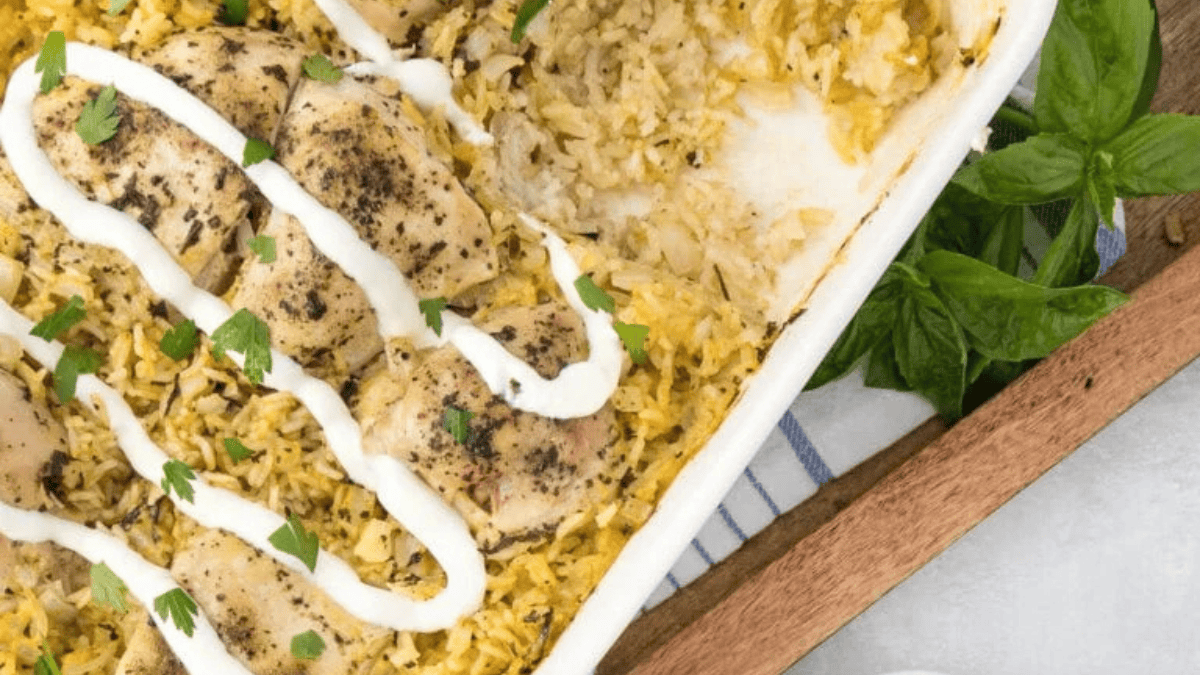 A casserole dish filled with baked chicken and yellow rice, inspired by classic chicken casserole recipes, topped with white sauce and fresh herbs. The dish sits on a wooden tray next to a bunch of fresh basil.