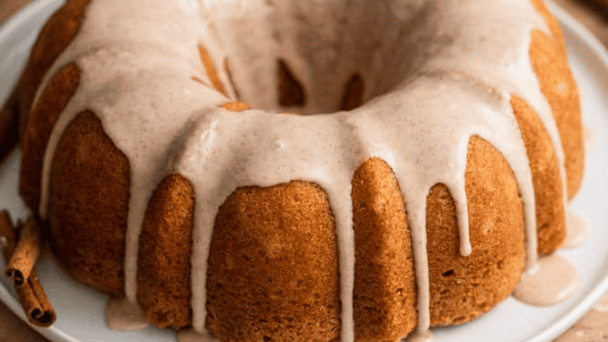 A round bundt cake with a light brown glaze drizzled over the top, pooling slightly at the sides, sits on a white plate. Cinnamon sticks are placed beside the cake, making it a perfect choice for lovers of cinnamon desserts.