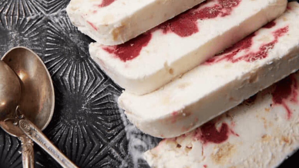 Four slices of a creamy white dessert with red fruit swirls are arranged on a patterned dark plate next to two vintage silver spoons, making it a festive addition to your collection of white Christmas recipes.