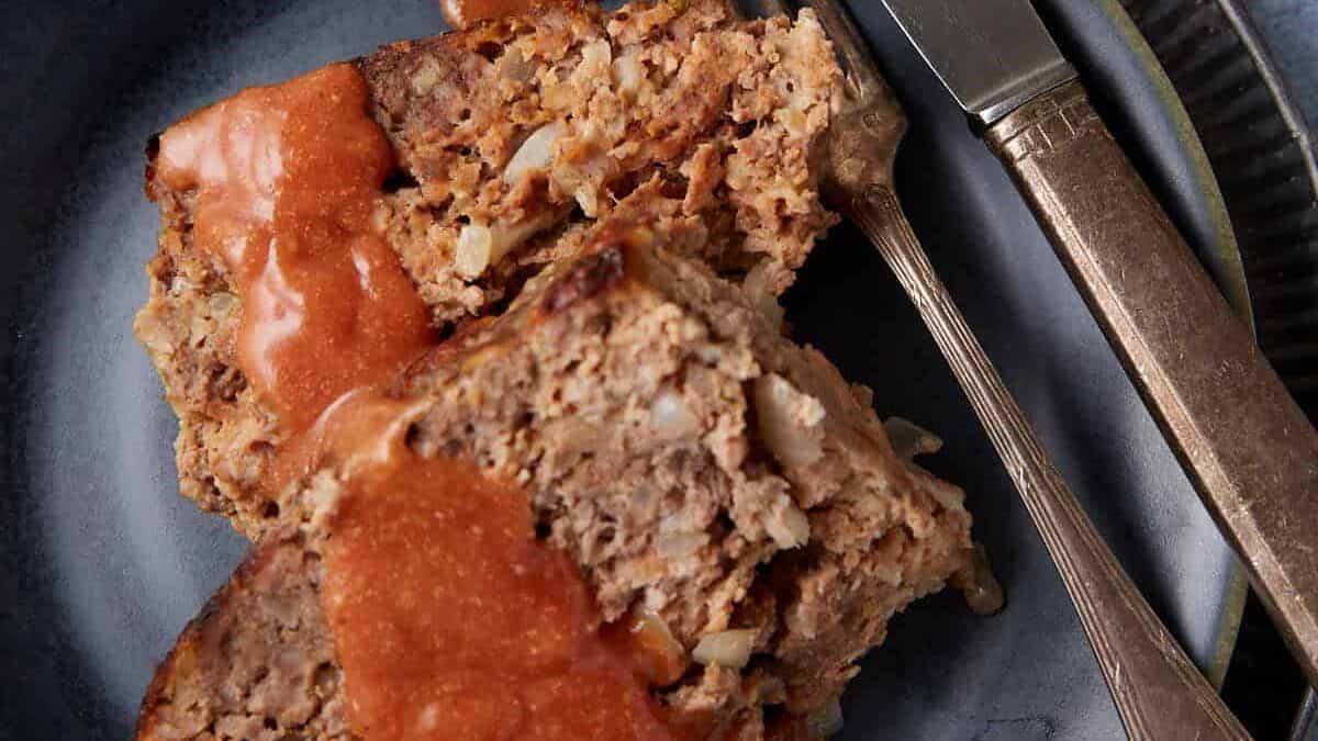 A close-up of a plate with slices of meatloaf, topped with a tomato-based sauce. The meatloaf is garnished with visible onion bits and is served with a fork and knife placed beside it. The plate is set on a textured surface and is part of a rustic table setting.