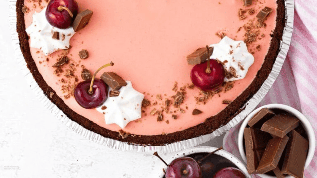 A pink mousse no bake pie in a chocolate crust, topped with cherries, whipped cream swirls, and chocolate shavings. Pieces of chocolate and cherries are in bowls nearby, with a pink-striped cloth at the side.