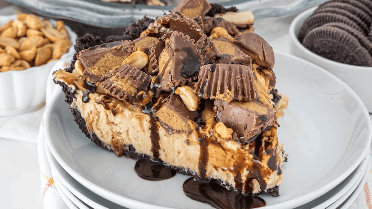 A slice of peanut butter pie with a chocolate cookie crust, perfect for fans of chocolate desserts, is topped with chopped peanut butter cups, peanuts, and drizzled with chocolate syrup on a white plate.