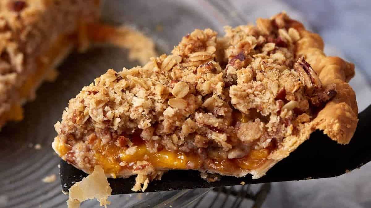 A slice of pie with a crumbly streusel topping is being lifted from a glass pie dish. The pie filling, visible from the cut slice, appears to be a rich, juicy peach mixture. A whole peach is in the blurred background on a light blue surface.