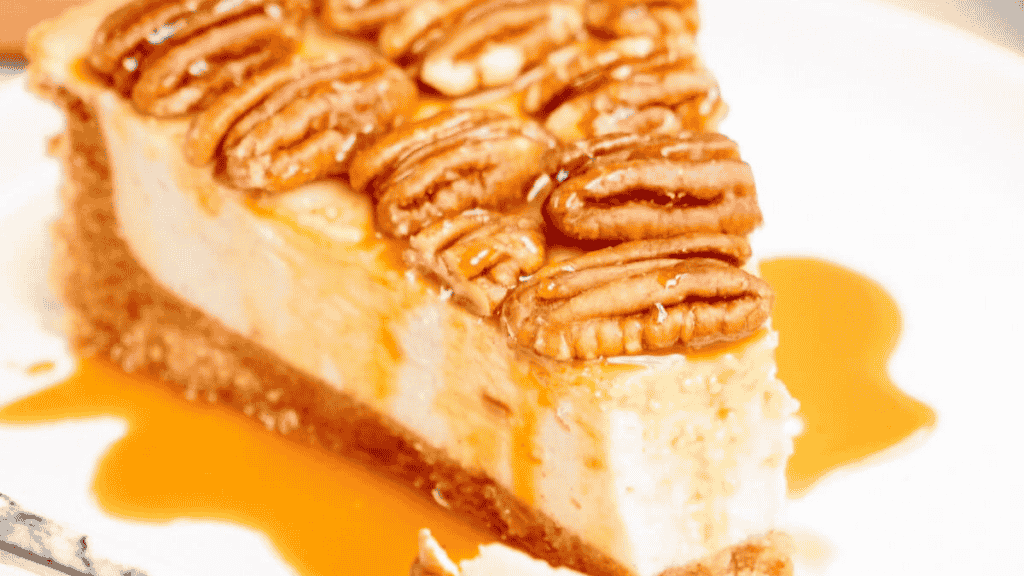 A slice of cheesecake topped with whole pecans and drizzled with caramel sauce sits on a white plate—a delightful addition to any pecan desserts spread.
