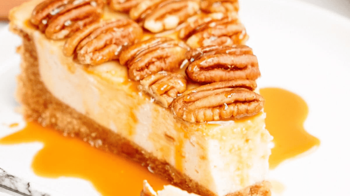A slice of cheesecake topped with whole pecans and drizzled with caramel sauce sits on a white plate—a delightful addition to any pecan desserts spread.