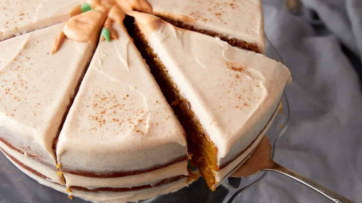 A frosted layer cake with light brown icing, decorated with a dusting of cinnamon and a piped frosting design—perfect for thanksgiving cake ideas. One slice is being lifted out with a cake server.