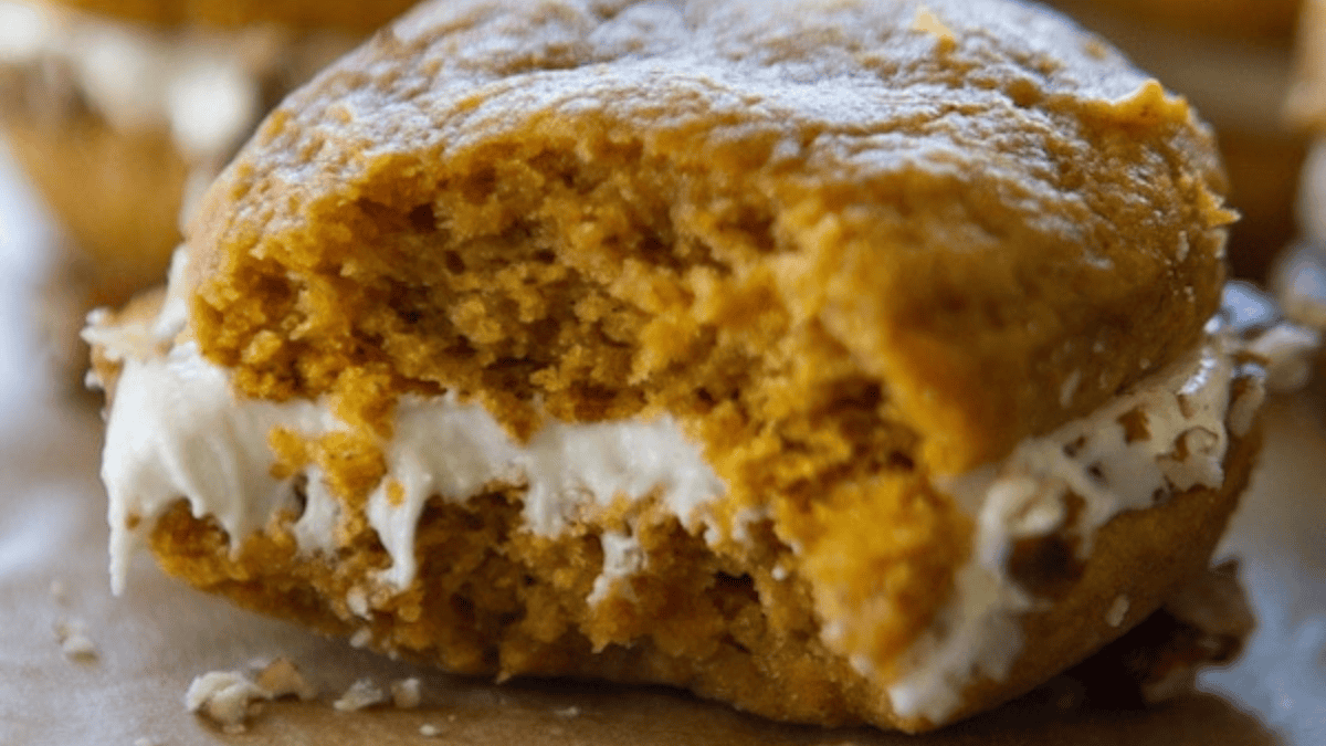A close-up of a pumpkin whoopie pie with a bite taken out, showing creamy white filling between two soft, orange cookies—a delightful twist on classic pumpkin desserts.